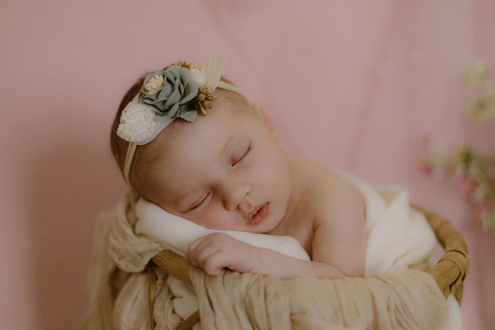 A sleeping newborn swaddled in beige fabric wearing a delicate green floral headband rests on a pink background.