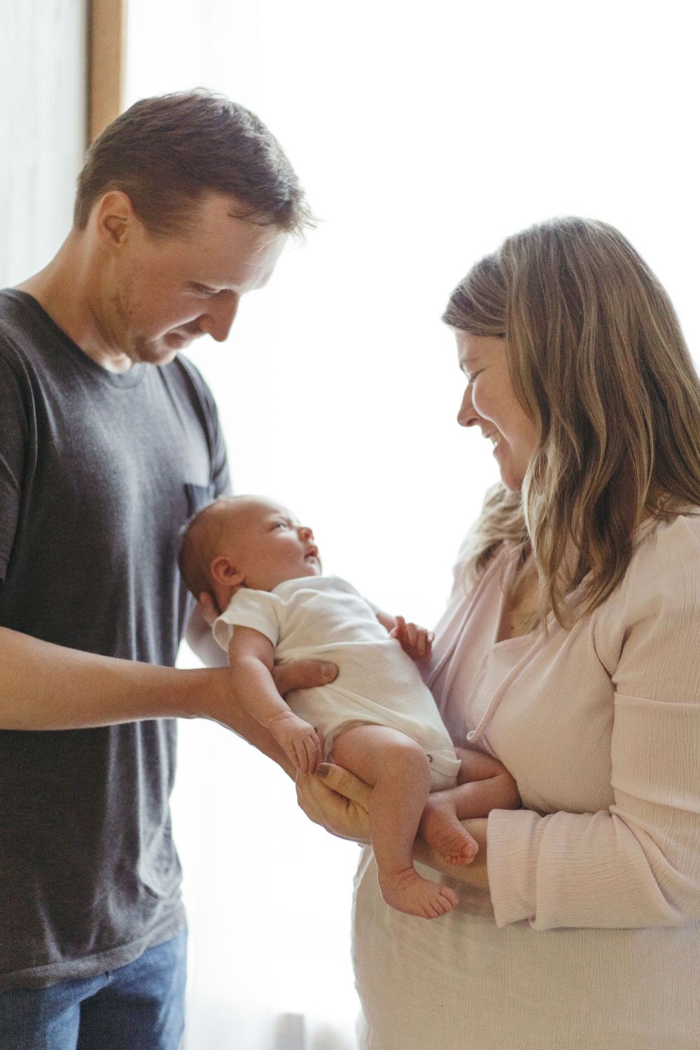 Loving parents tenderly hold and gaze at their newborn baby in a bright, intimate indoor setting.