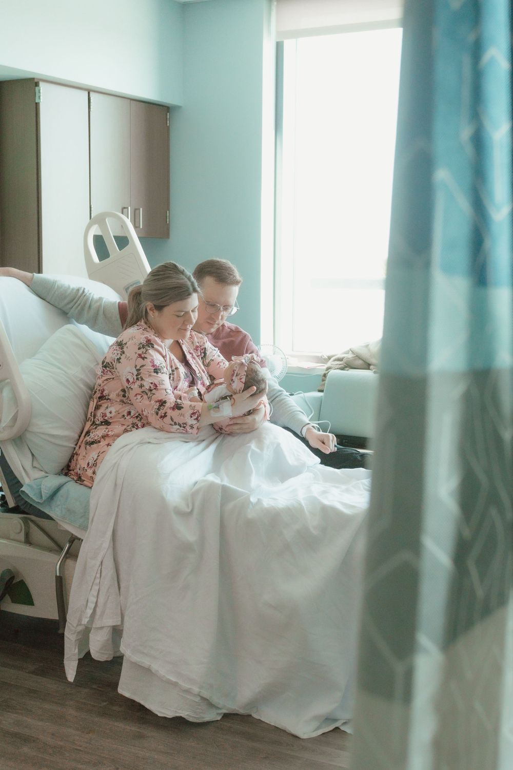 Parent in floral robe embraces newborn baby while reclining on white bedding in bright hospital room.