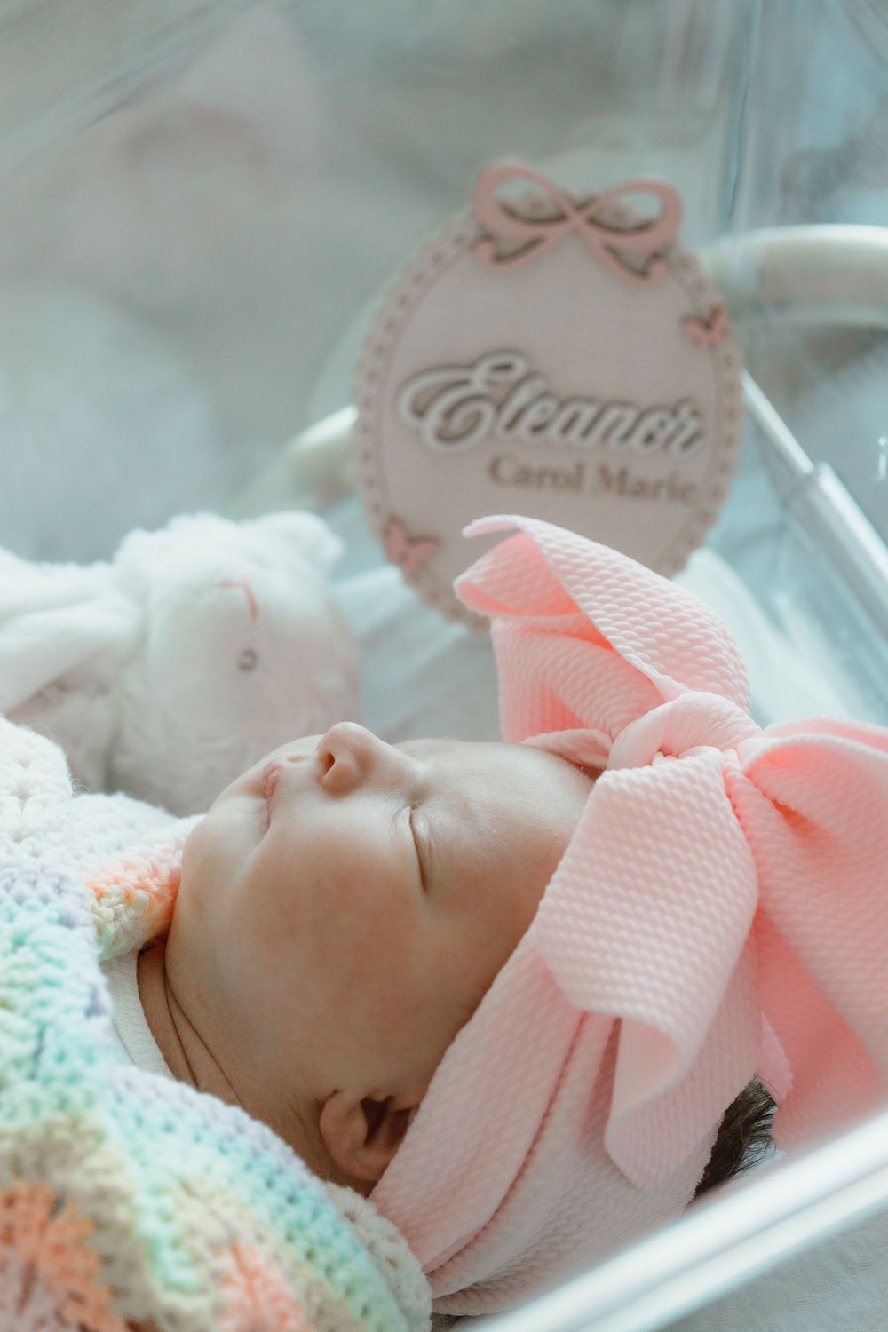 A sleeping newborn baby in a pink sweater lies in a bassinet with a pastel rainbow blanket and personalized name tag.