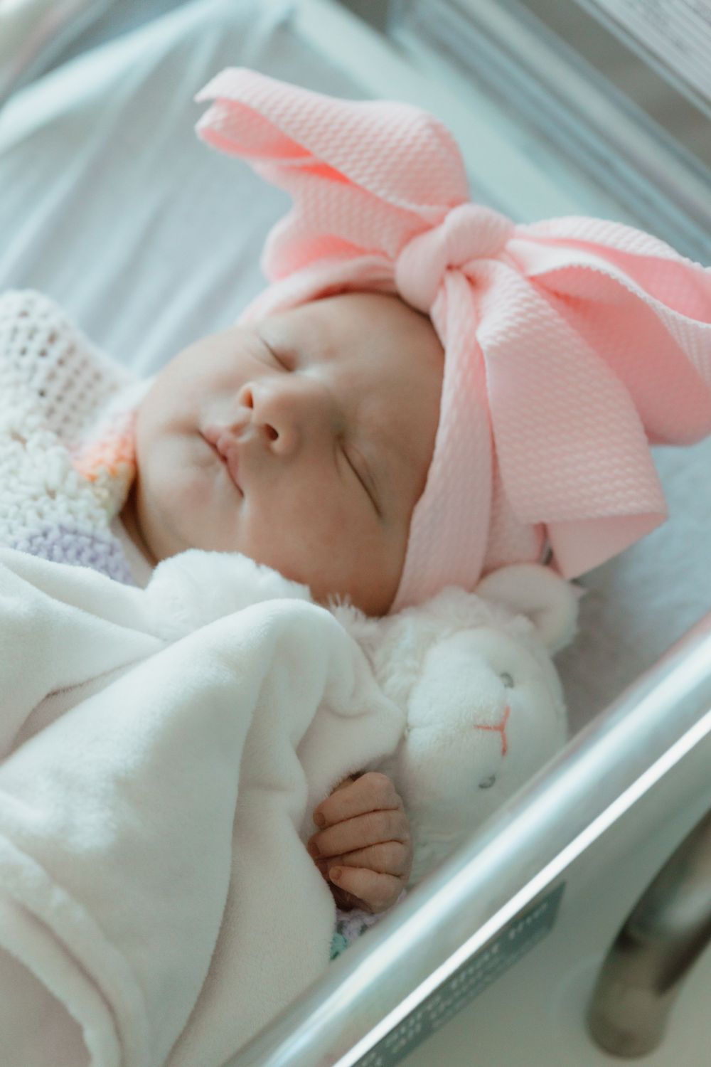 A sleeping newborn wearing a pink bow hat and white blanket rests in a hospital bassinet.