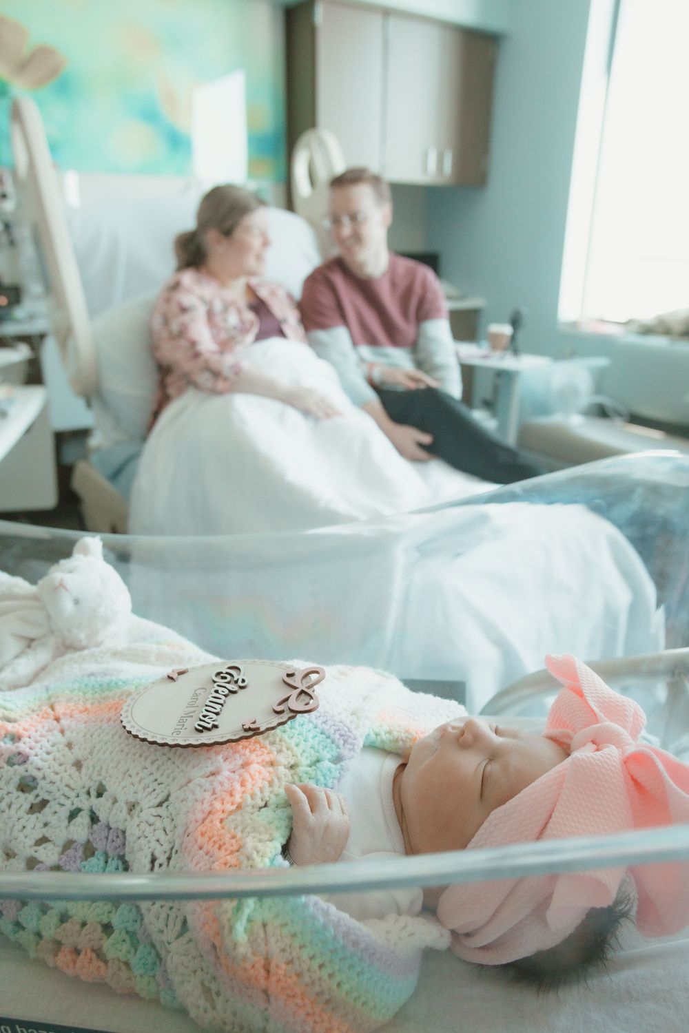 A newborn baby in a pink bow sleeps in a hospital bassinet with family in the background.