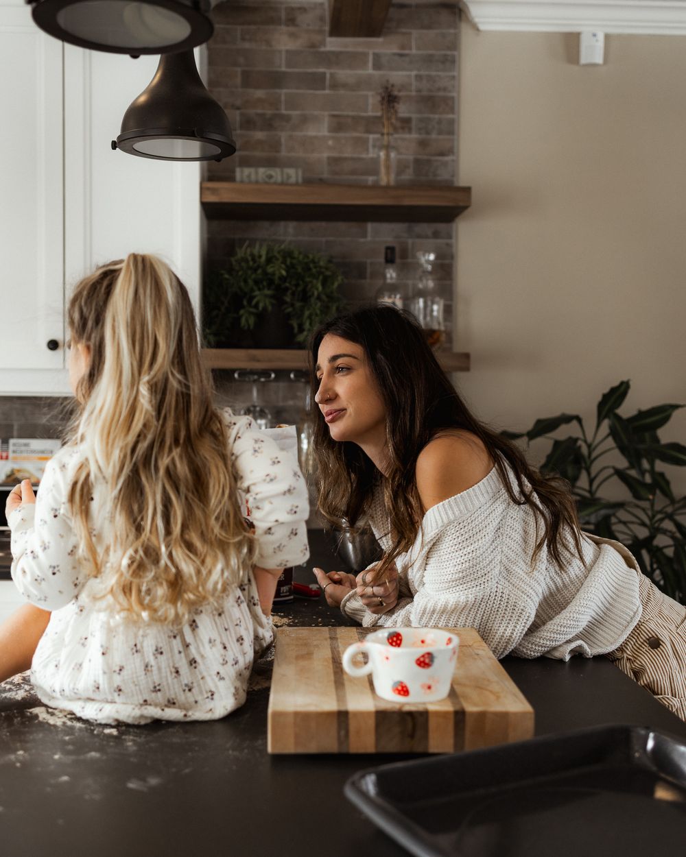 Two friends laughing and enjoying coffee together at a kitchen counter with exposed brick wall.