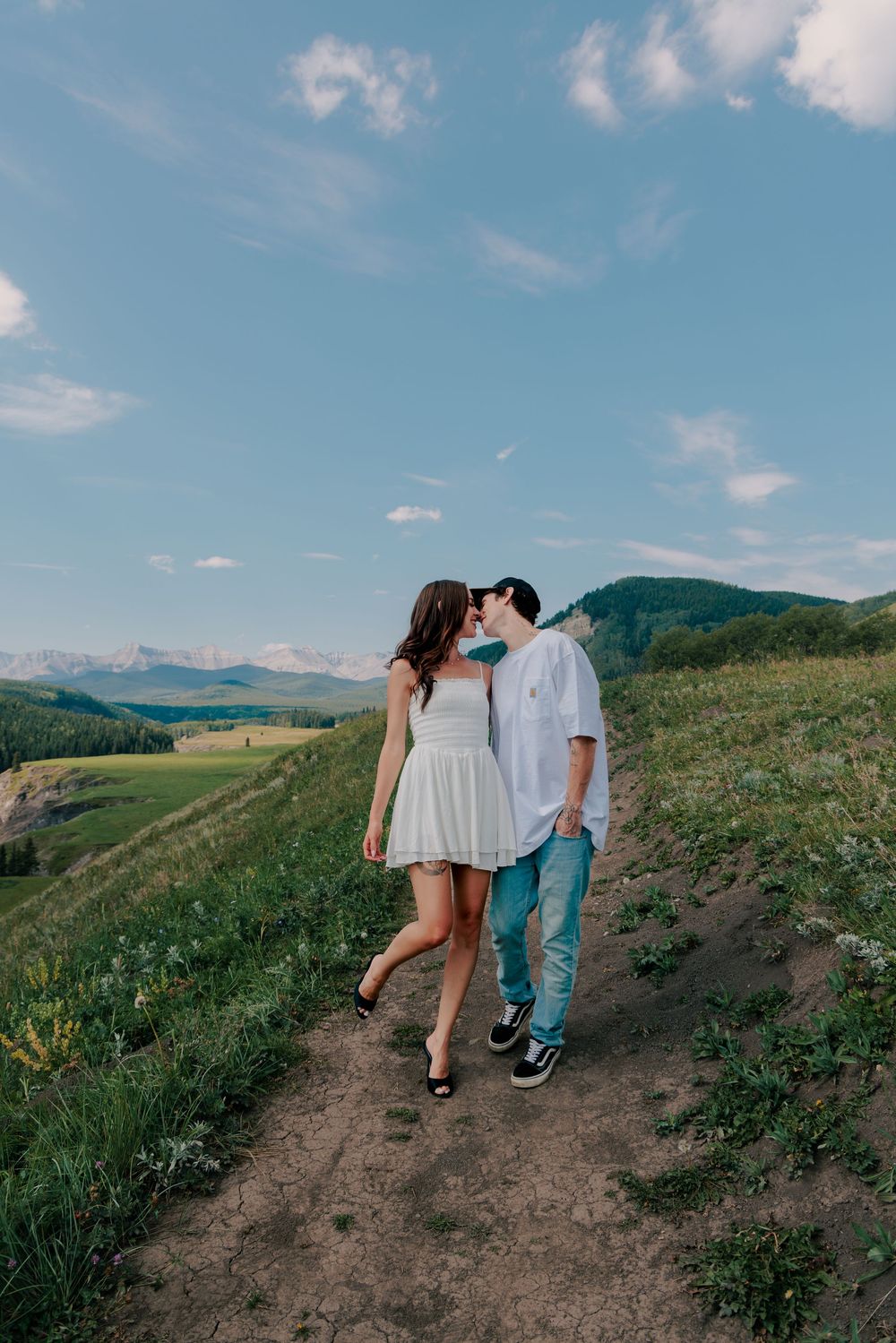 A couple walks hand in hand along a scenic mountain trail under blue skies.