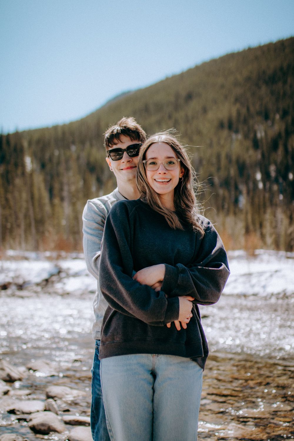 Two people embrace and laugh together in a winter mountain setting with snow on the ground.