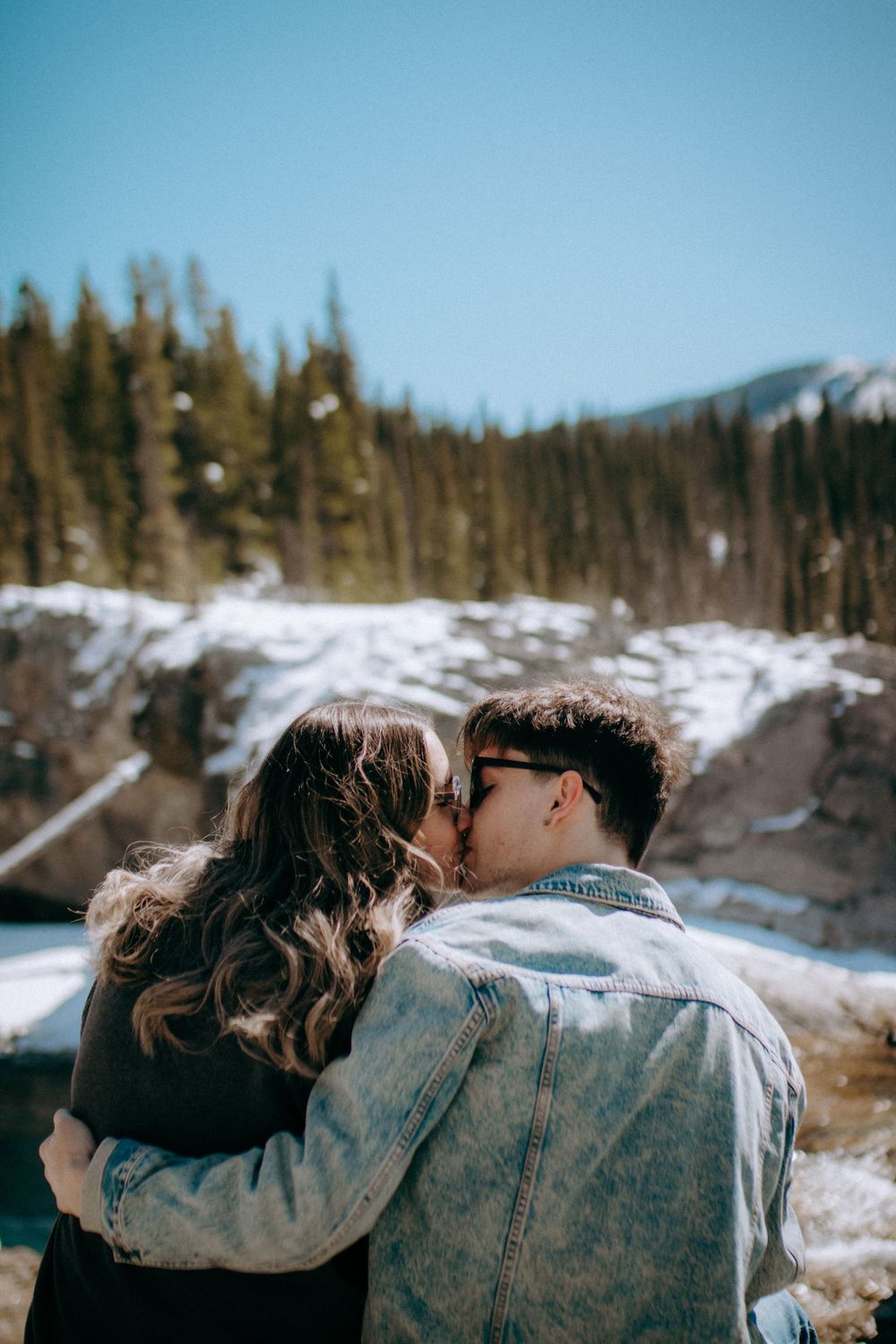 Couple shares intimate moment against snowy mountain backdrop and evergreen forest.