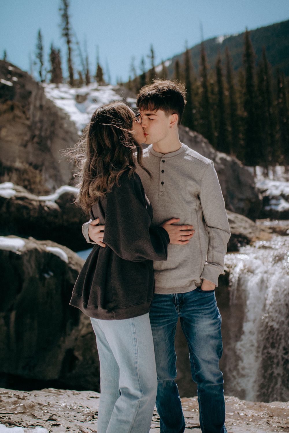 Two people share an intimate moment near a mountain waterfall in winter.