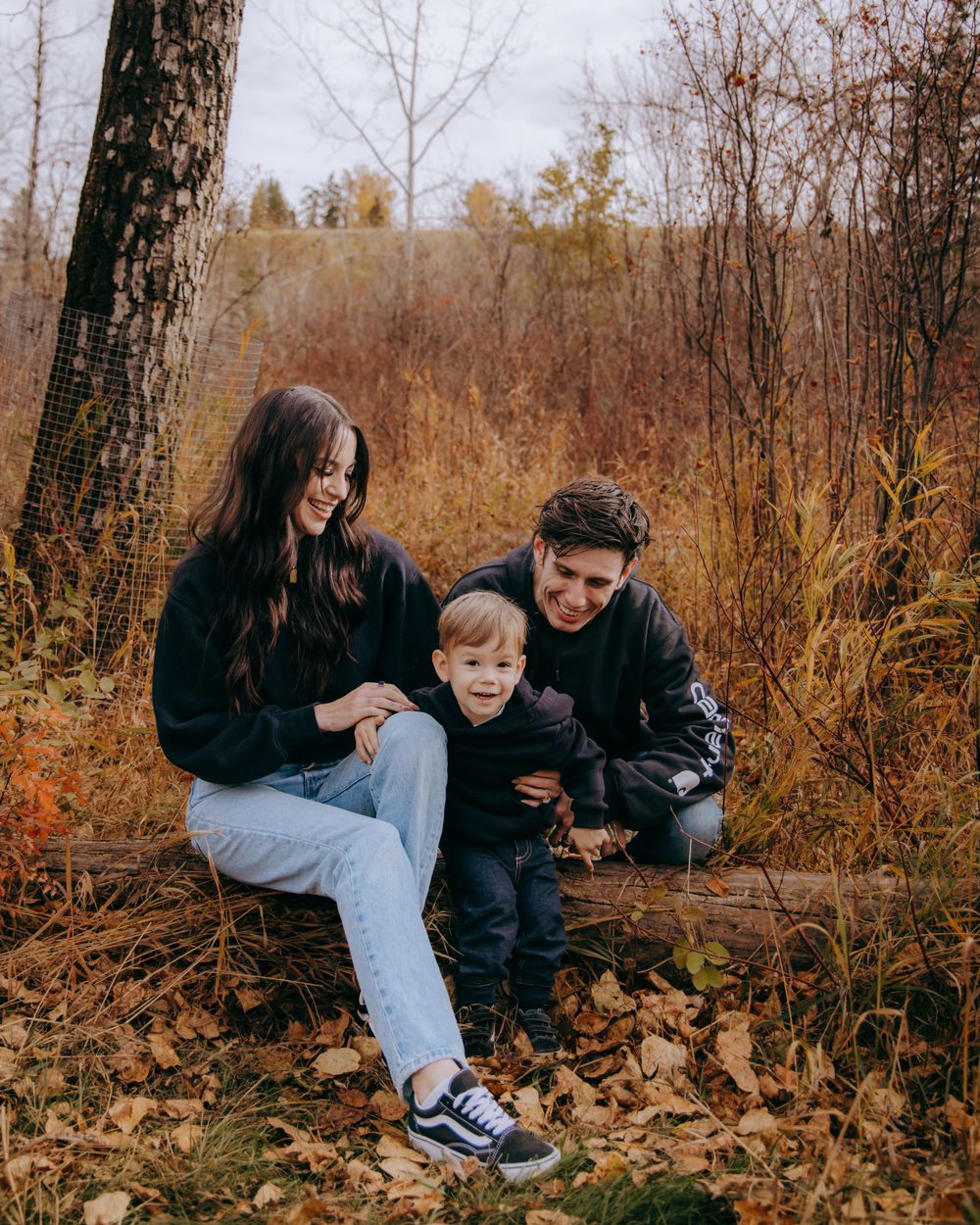 People play in fallen autumn leaves beside a tree trunk in a natural setting.