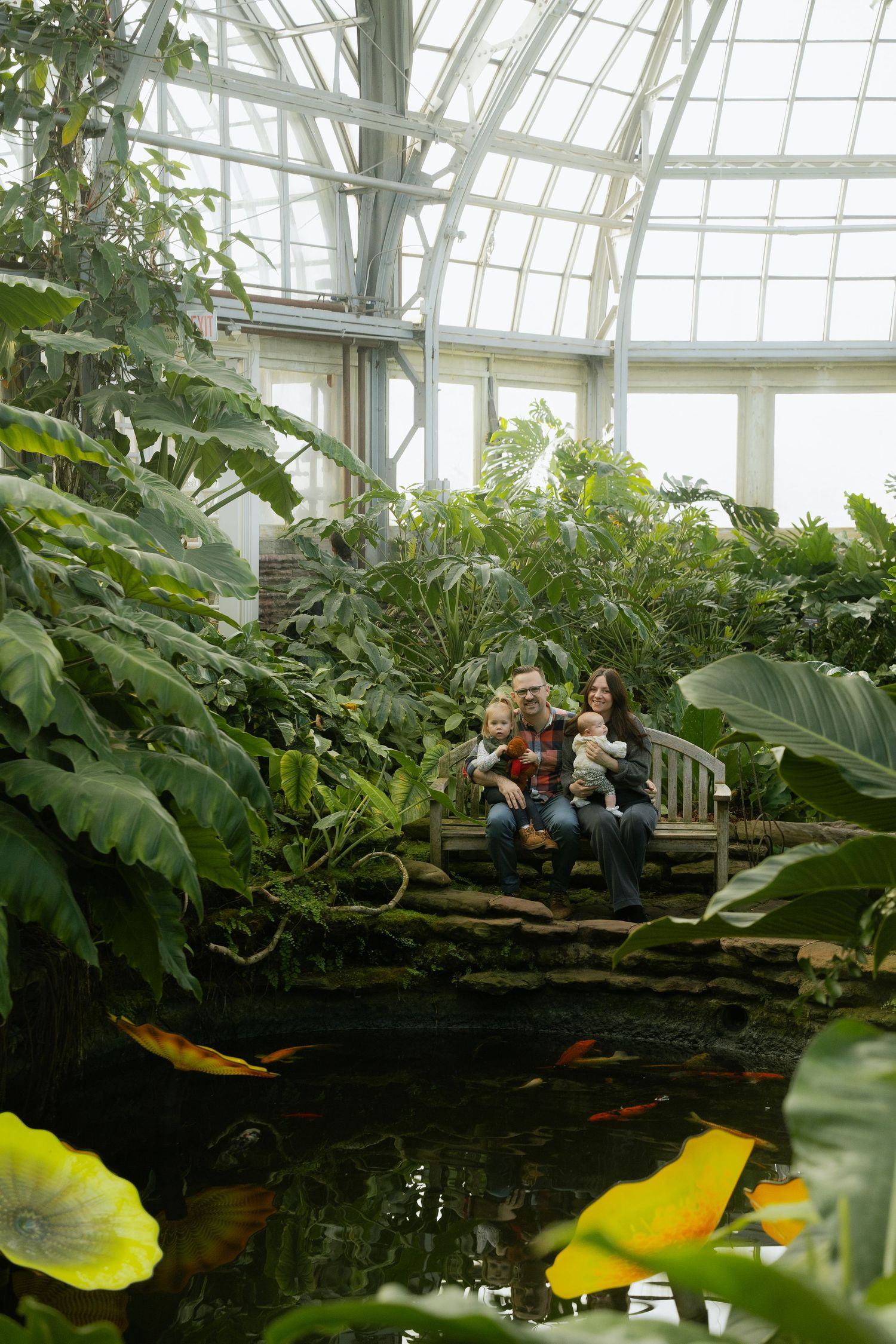 A greenhouse conservatory with lush tropical plants and a decorative pond featuring koi fish.