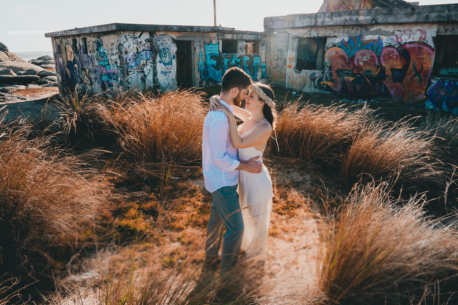 A romantic couple embraces among dried grass with urban graffiti walls in the background during sunset.