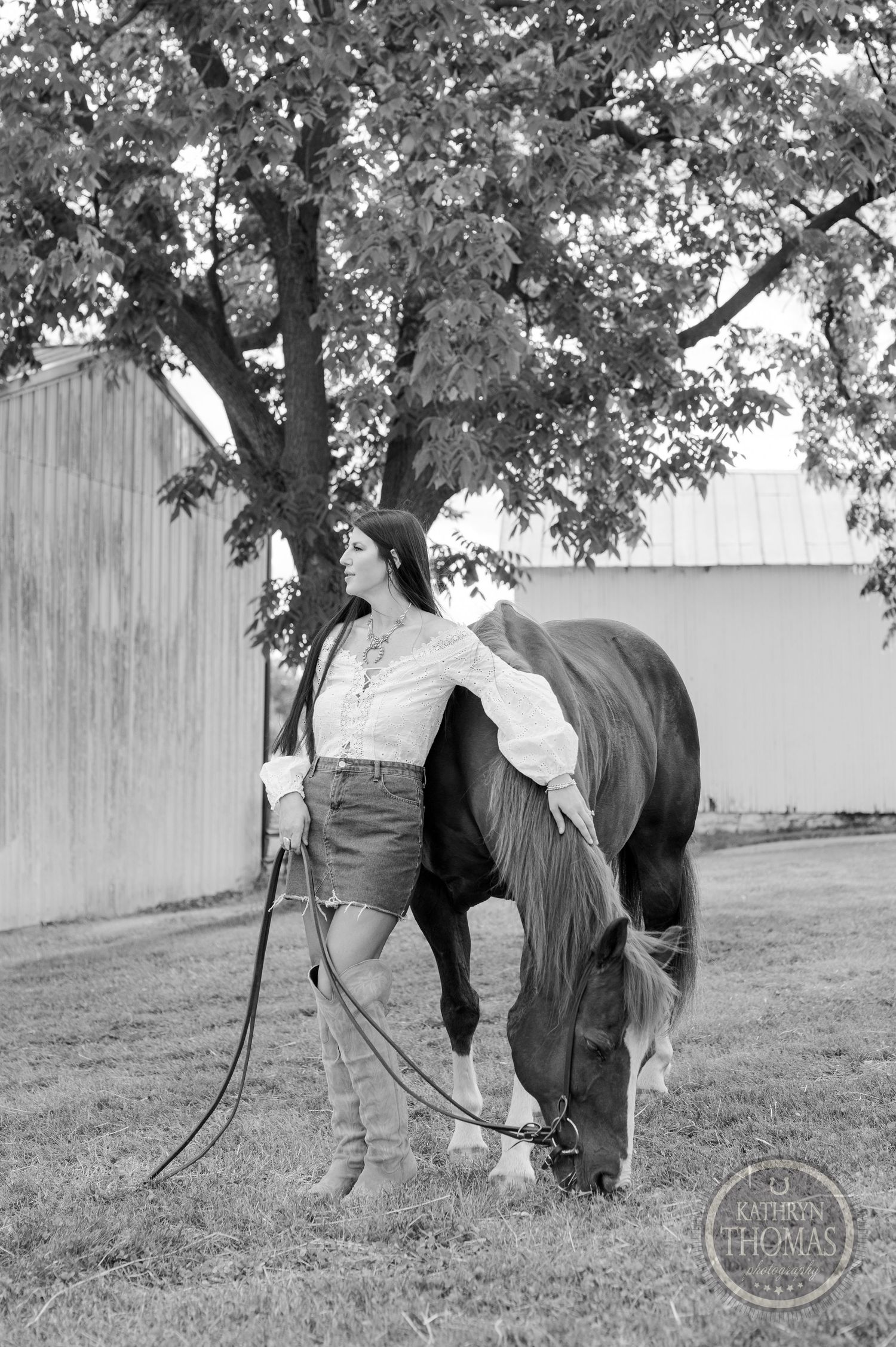 Black and white photo of woman holding reins while horse grazes near barn wall.