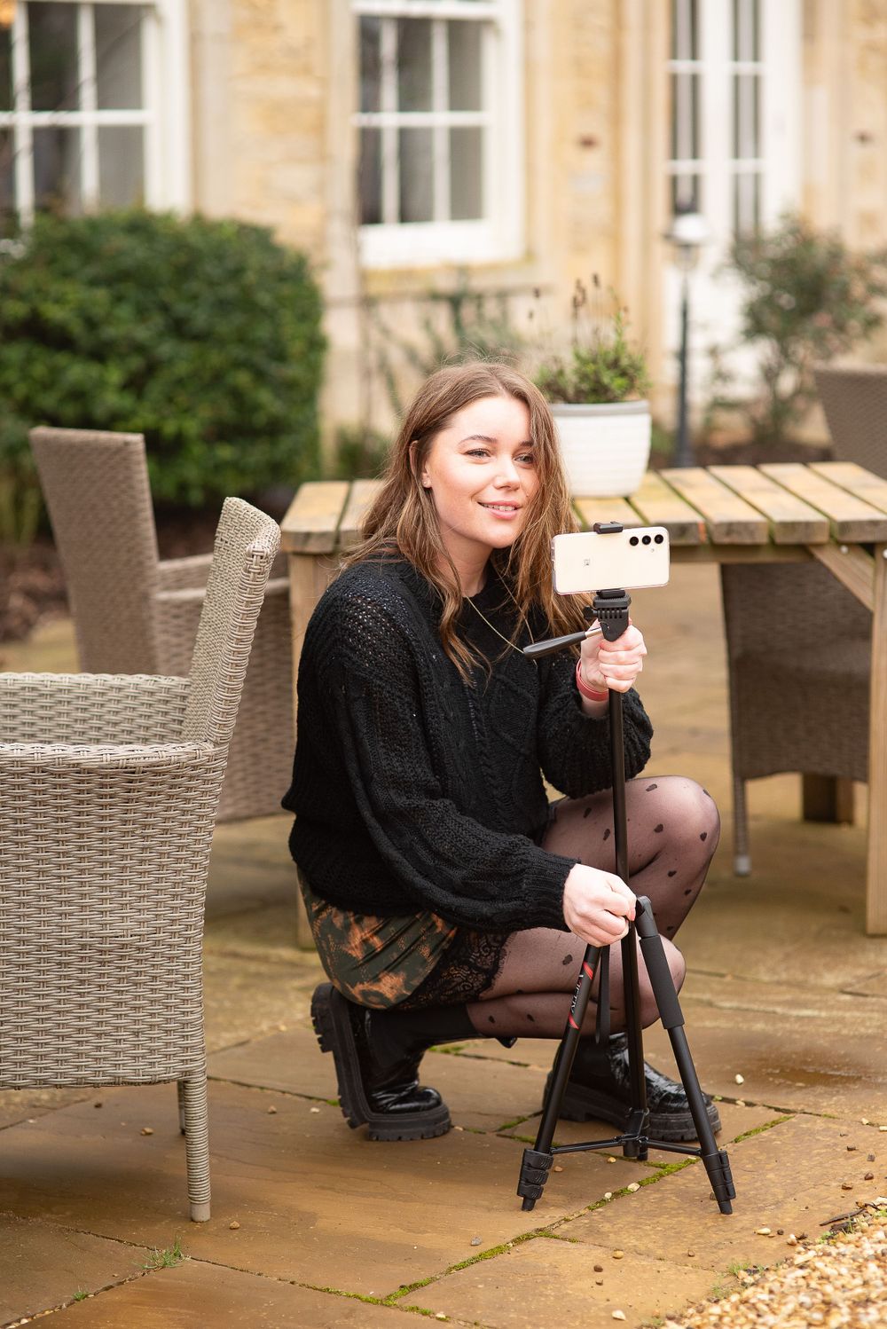 A photographer in a black sweater crouches with camera equipment on an outdoor stone patio with wicker furniture.