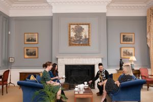 People relax in an elegant room with gray walls, blue armchairs, and a fireplace with framed artwork above it.