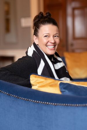 Smiling person in black coat and striped scarf relaxing on a blue couch with golden accents in a cozy interior.
