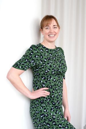 A person poses in a green leopard print dress with puff sleeves against a white curtain background.