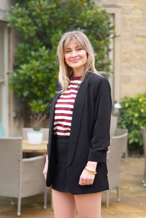 A person in a striped top and black blazer poses outdoors against stone building backdrop.