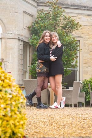 Two friends embrace while posing in stylish outfits on a gravel path outside an elegant stone building.