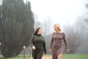 Two people in sweater dresses walk together on a misty path surrounded by evergreen trees on a foggy day.