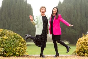 Two people posing playfully on a garden path surrounded by green cypress trees and yellow flowers.