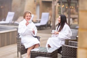 Two people in white spa robes relax and chat while sitting on wicker chairs on an outdoor patio.