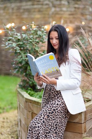 Woman in white blazer and leopard print skirt reads a book while sitting on wooden planter with twinkle lights in background.