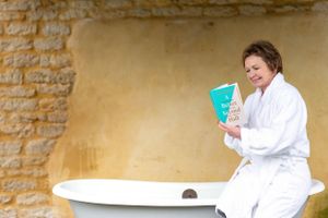 Someone wearing a white bathrobe reads a book while sitting on the edge of a vintage clawfoot bathtub against stone wall.