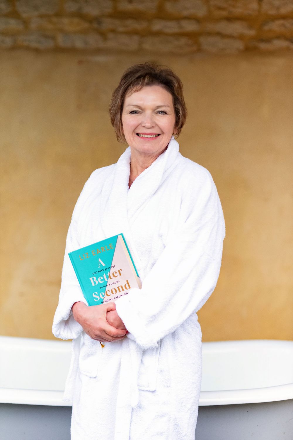 A person in a white bathrobe holds a blue book while posing against a beige wall near a bathtub.