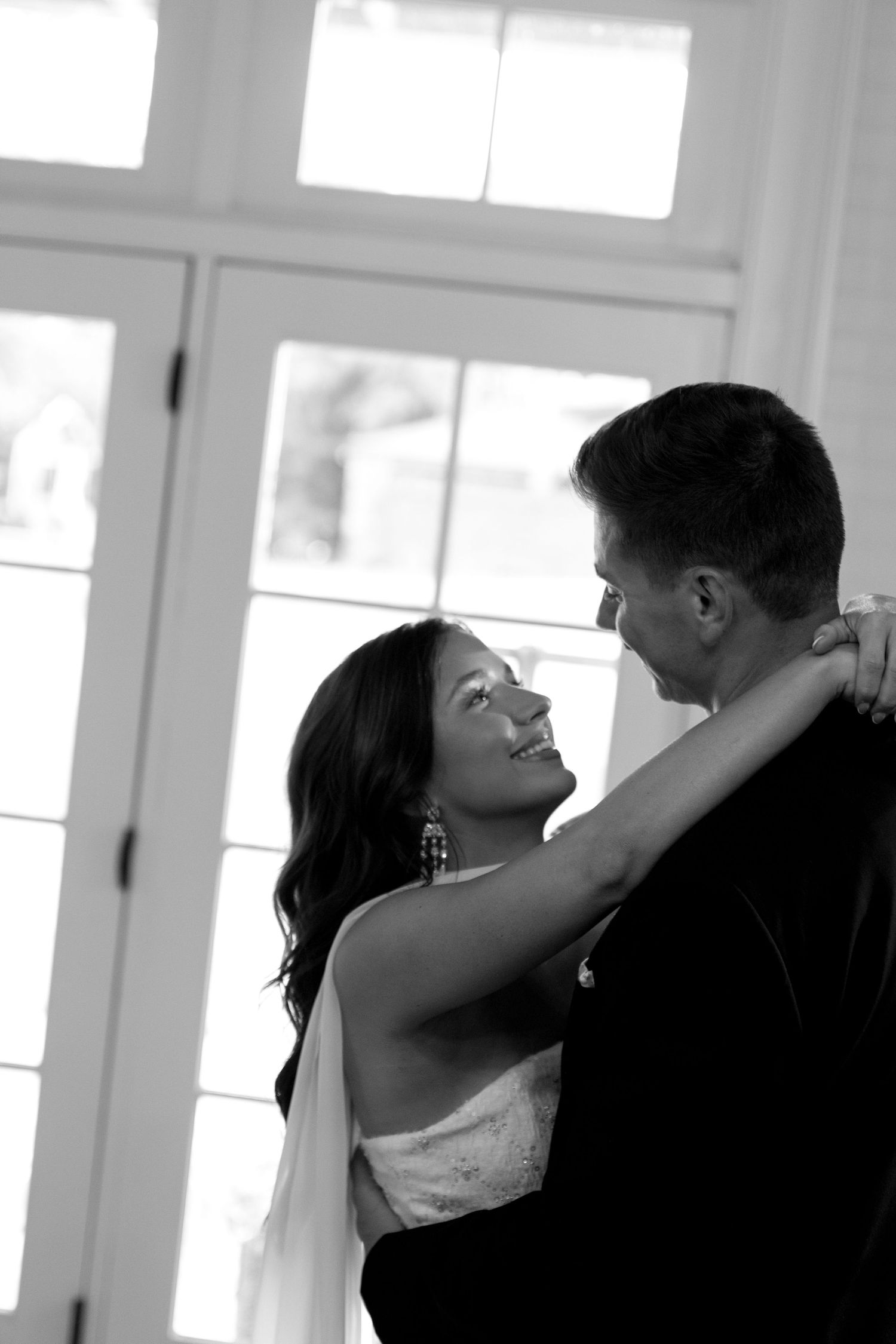 A couple shares a romantic dance in a sunlit room with white windows.