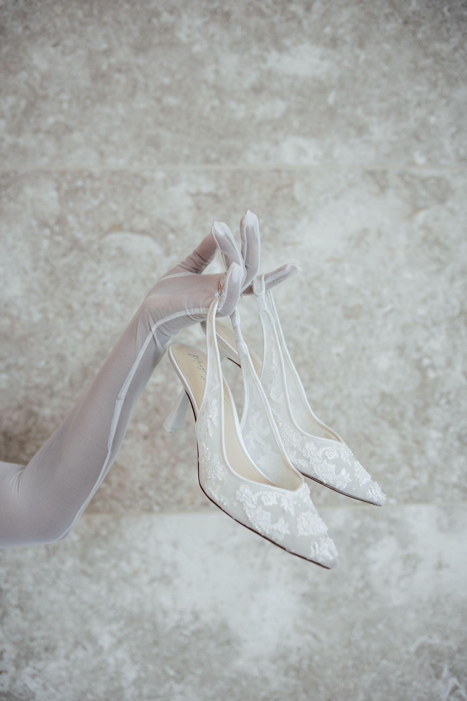 Delicate white lace bridal shoes arranged against a textured grey background.
