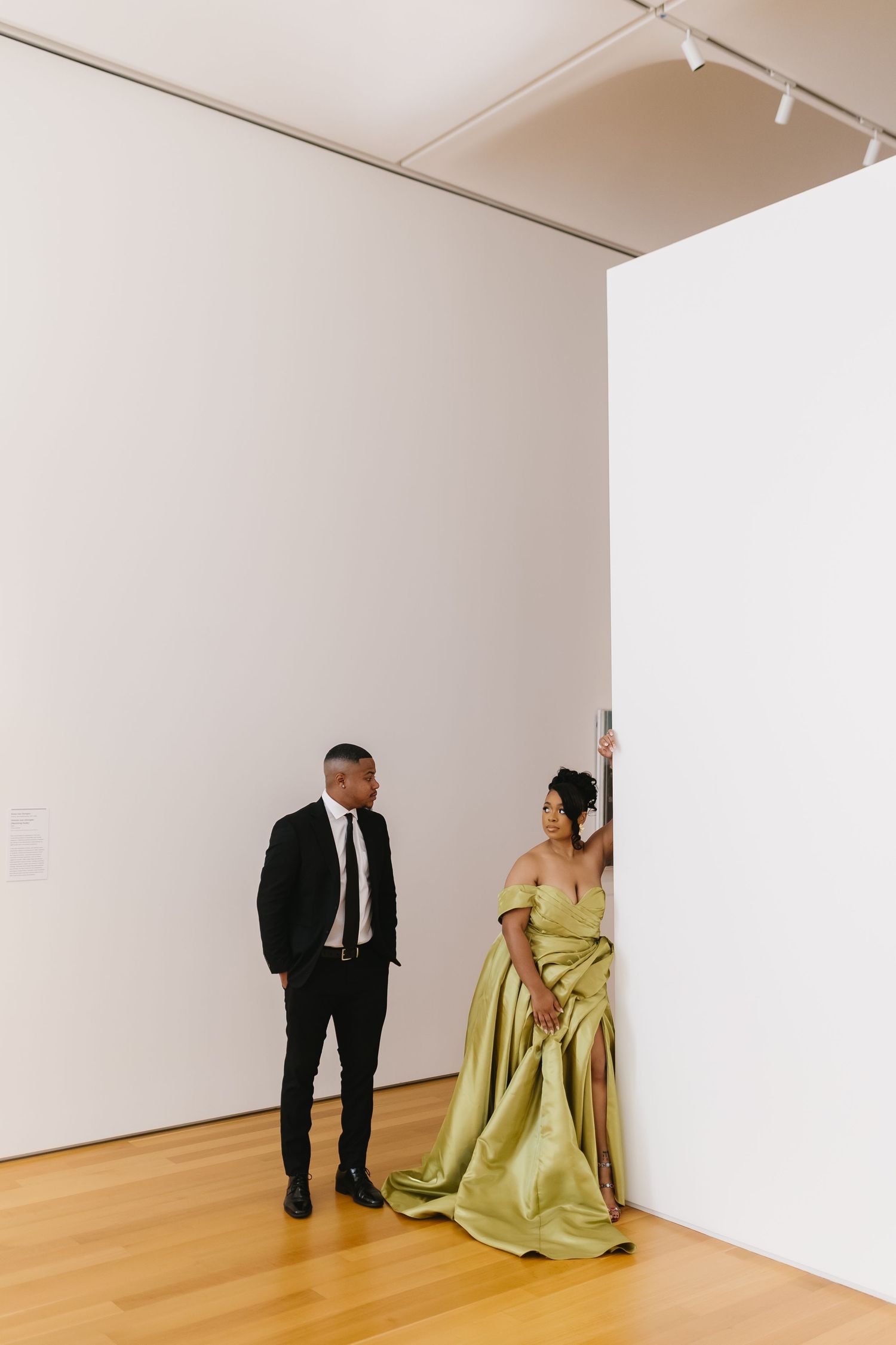 A couple poses against white gallery walls, with the woman wearing an elegant chartreuse gown.