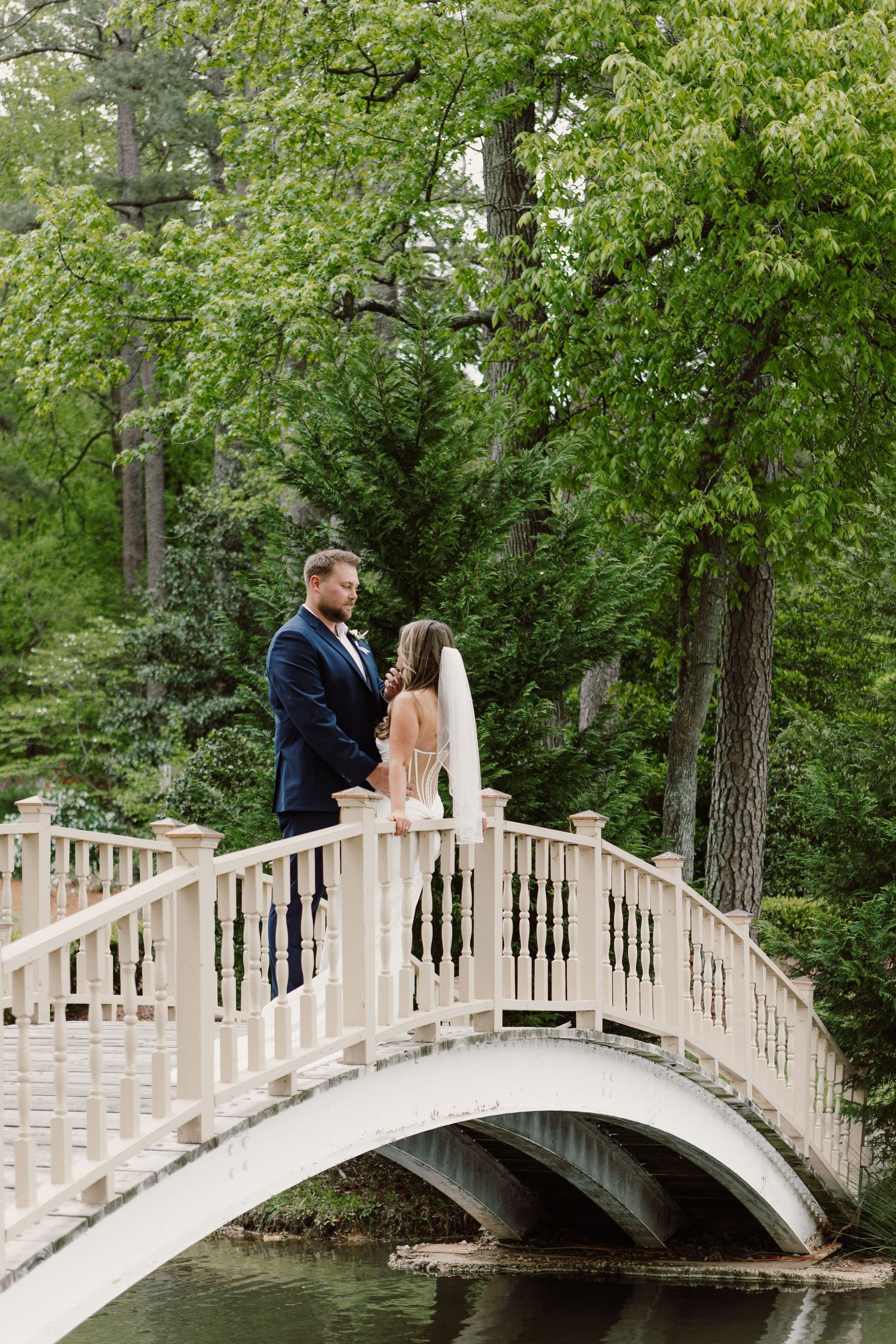 Romantic wedding couple standing on a white arched bridge surrounded by lush green trees.