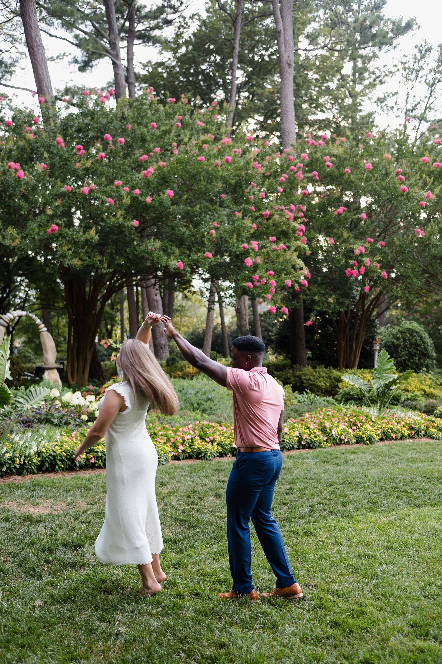 People celebrate joyfully in a garden with blooming pink flowers and pine trees.