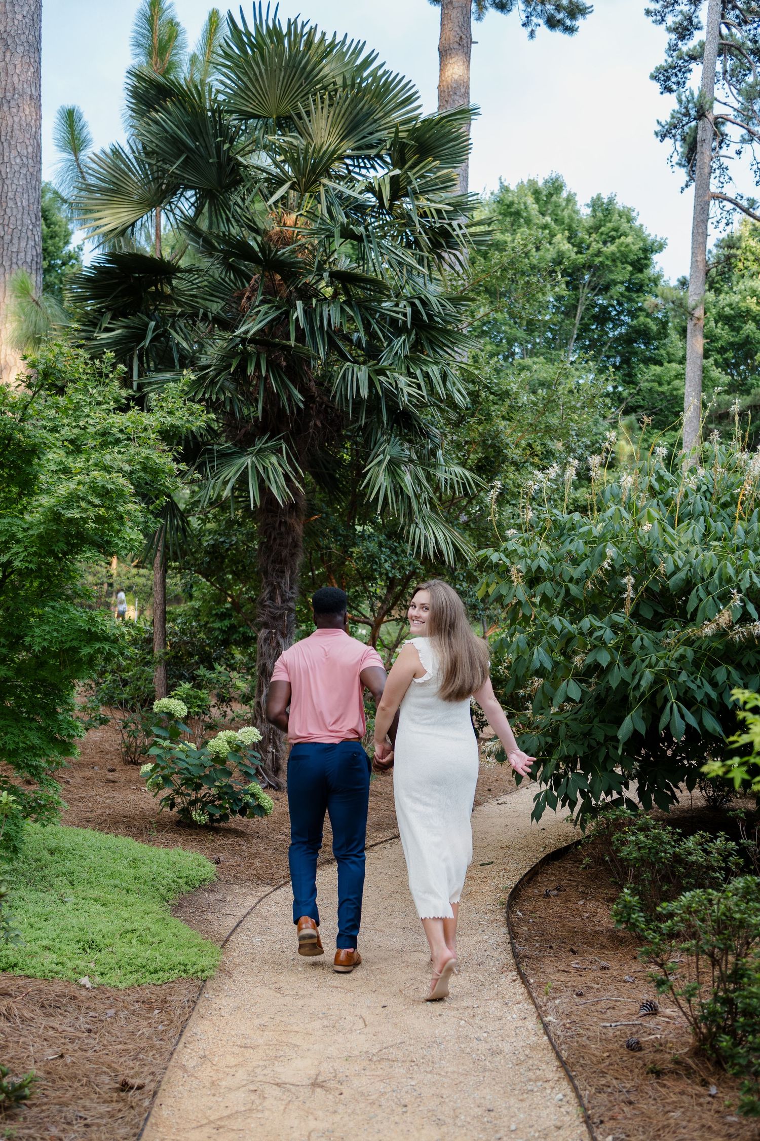 Two people walk along a winding path through a tropical garden with palm trees.