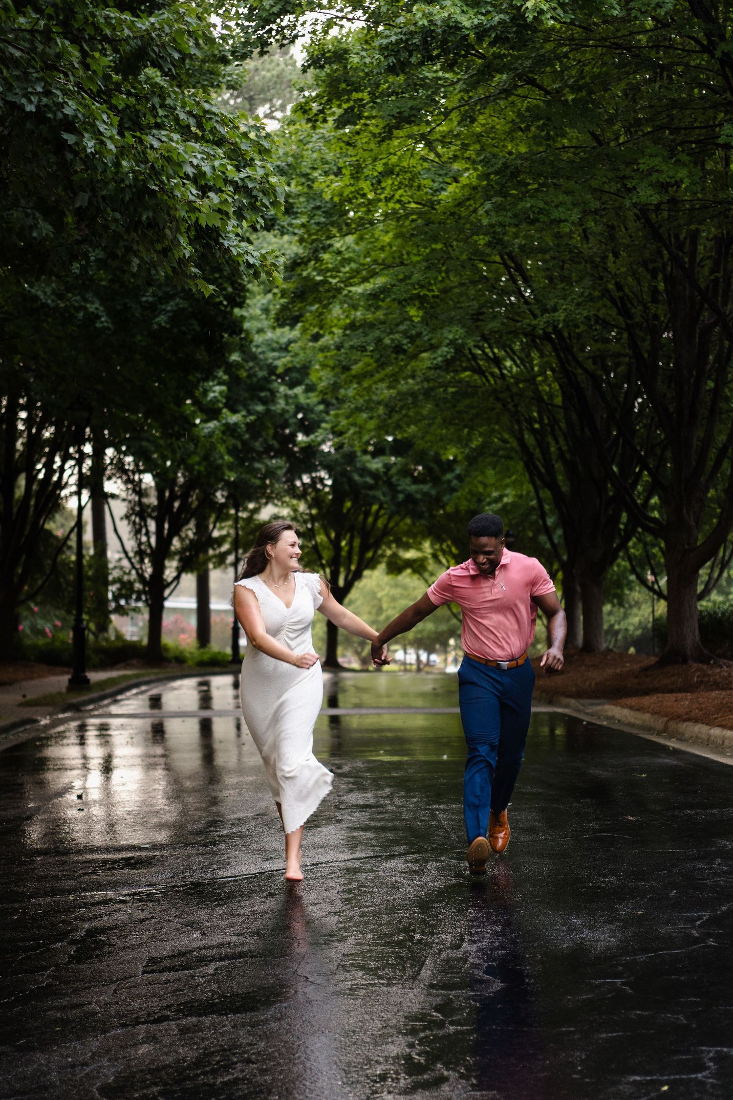 A couple joyfully runs together on a wet tree-lined path during a rainy day.