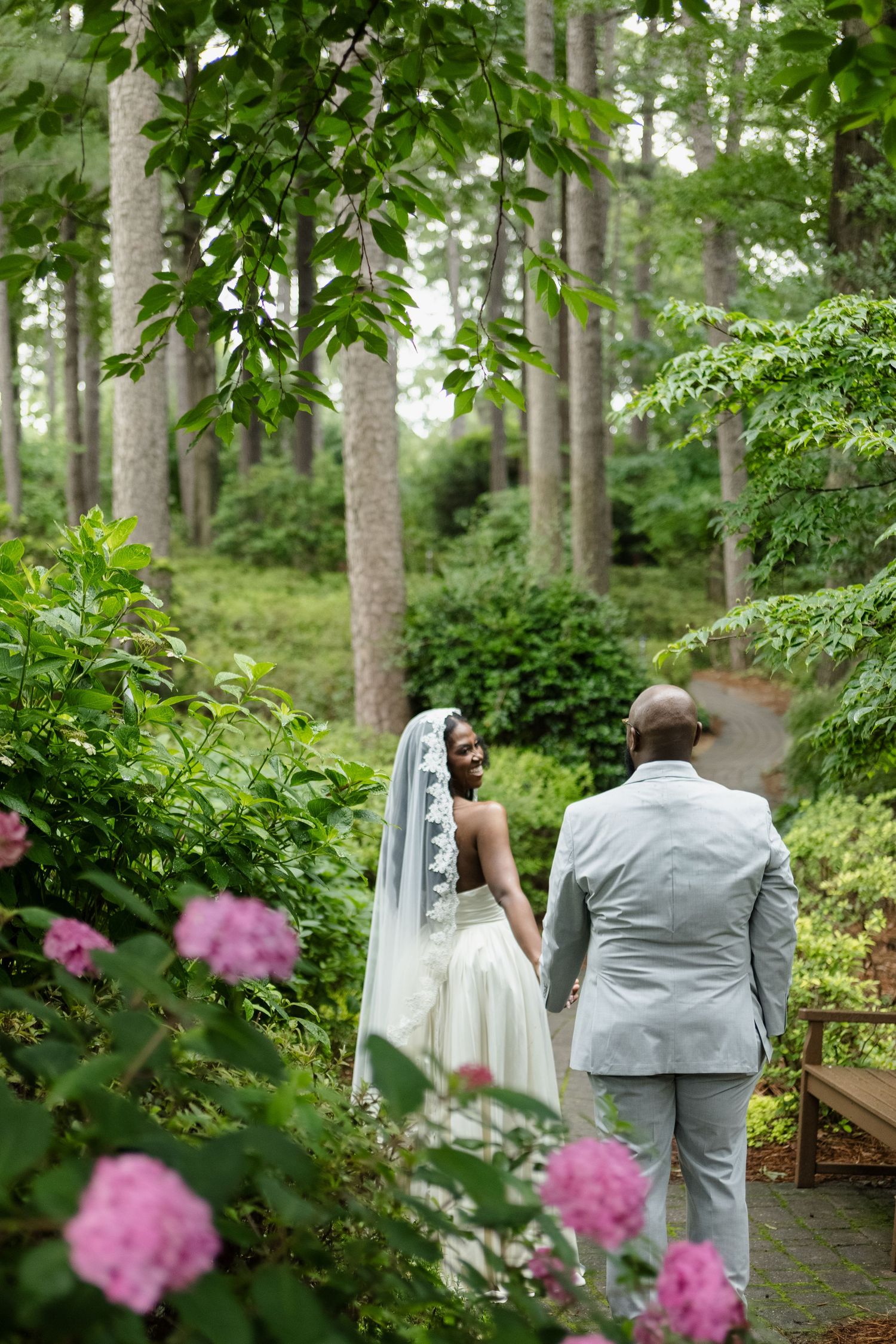 A couple walks down a garden path surrounded by pink flowers and tall trees.