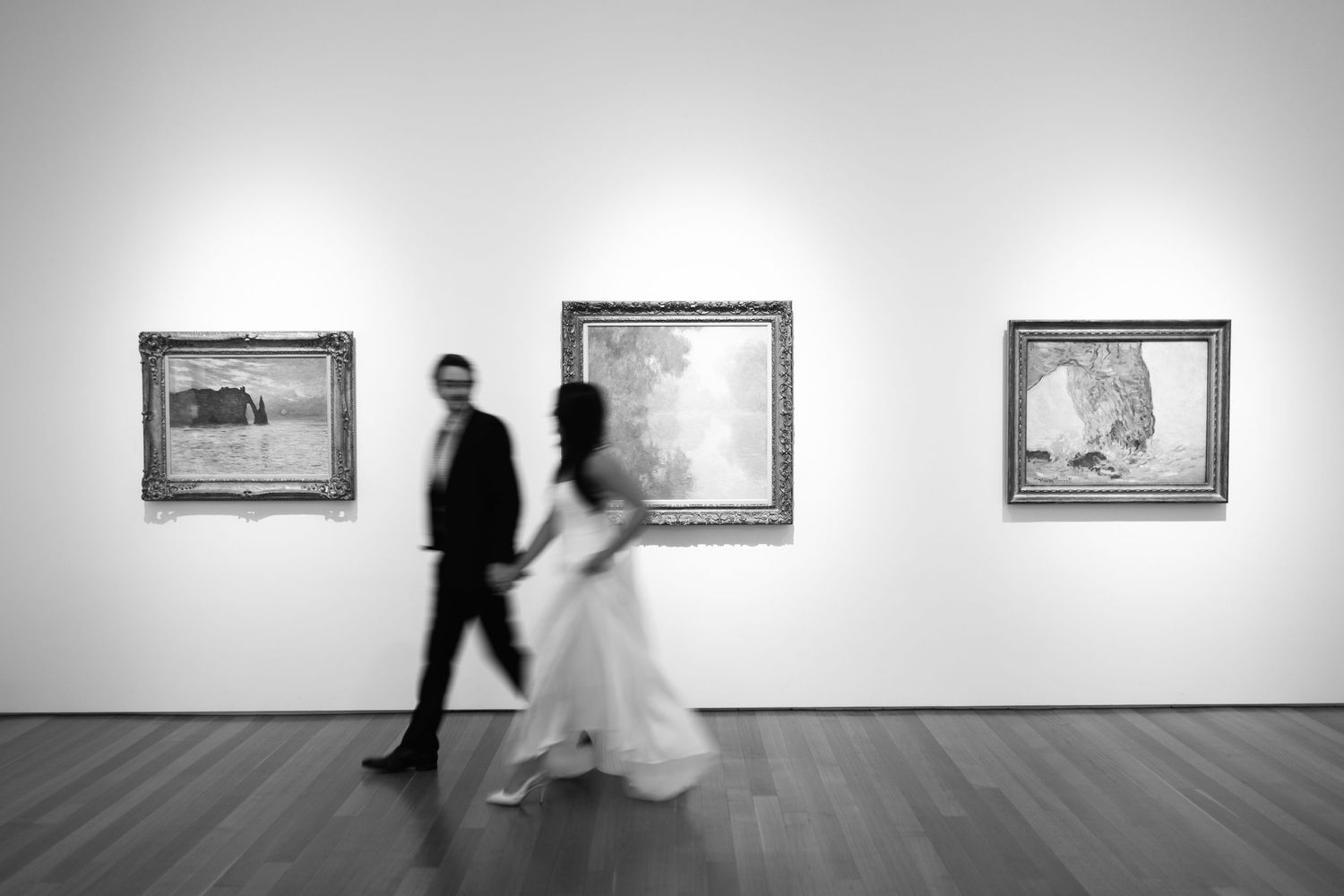 Black and white gallery scene with blurred figures walking past framed artwork.