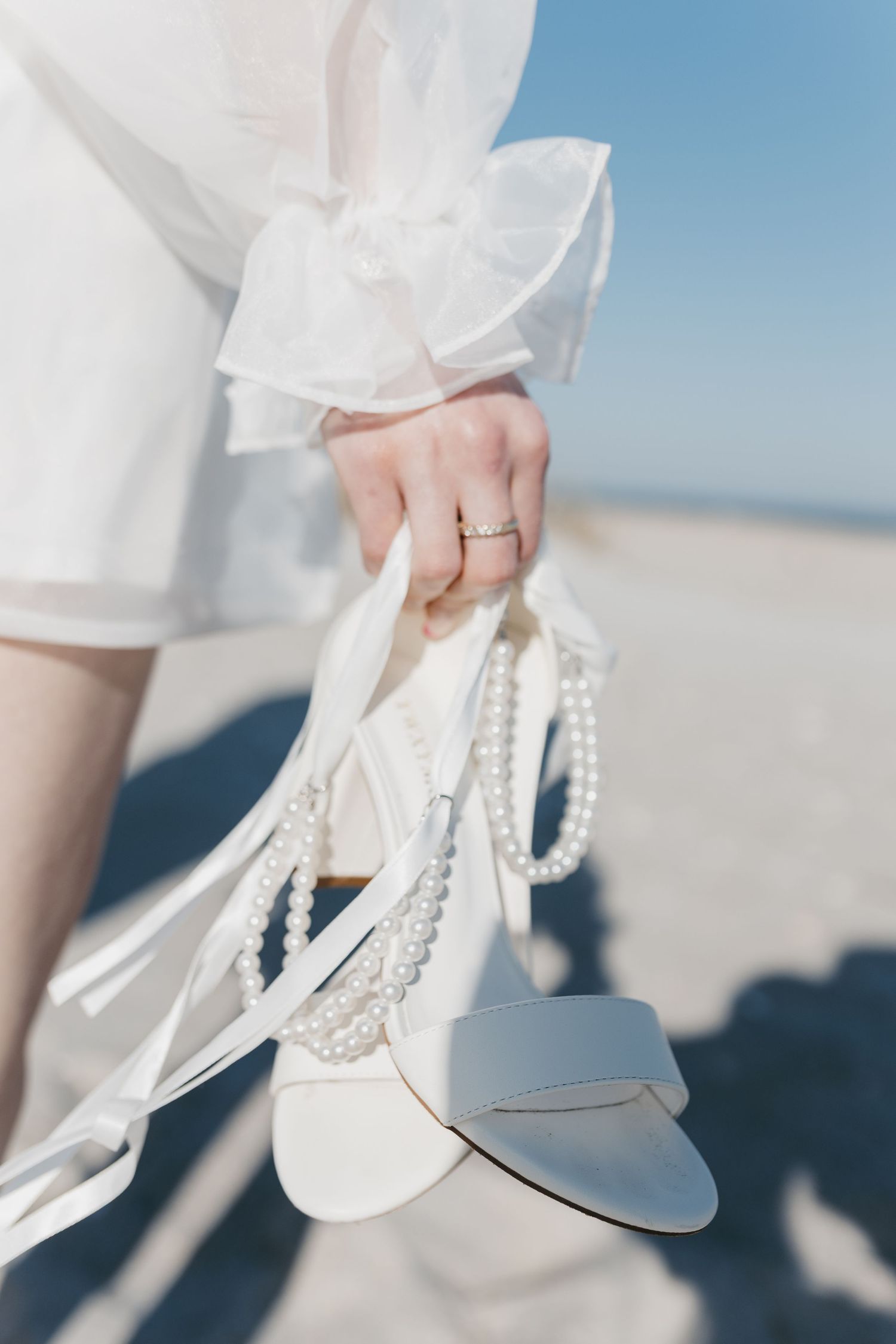 Elegant white bridal shoes with pearl details held by a hand wearing a wedding ring against a beach backdrop.