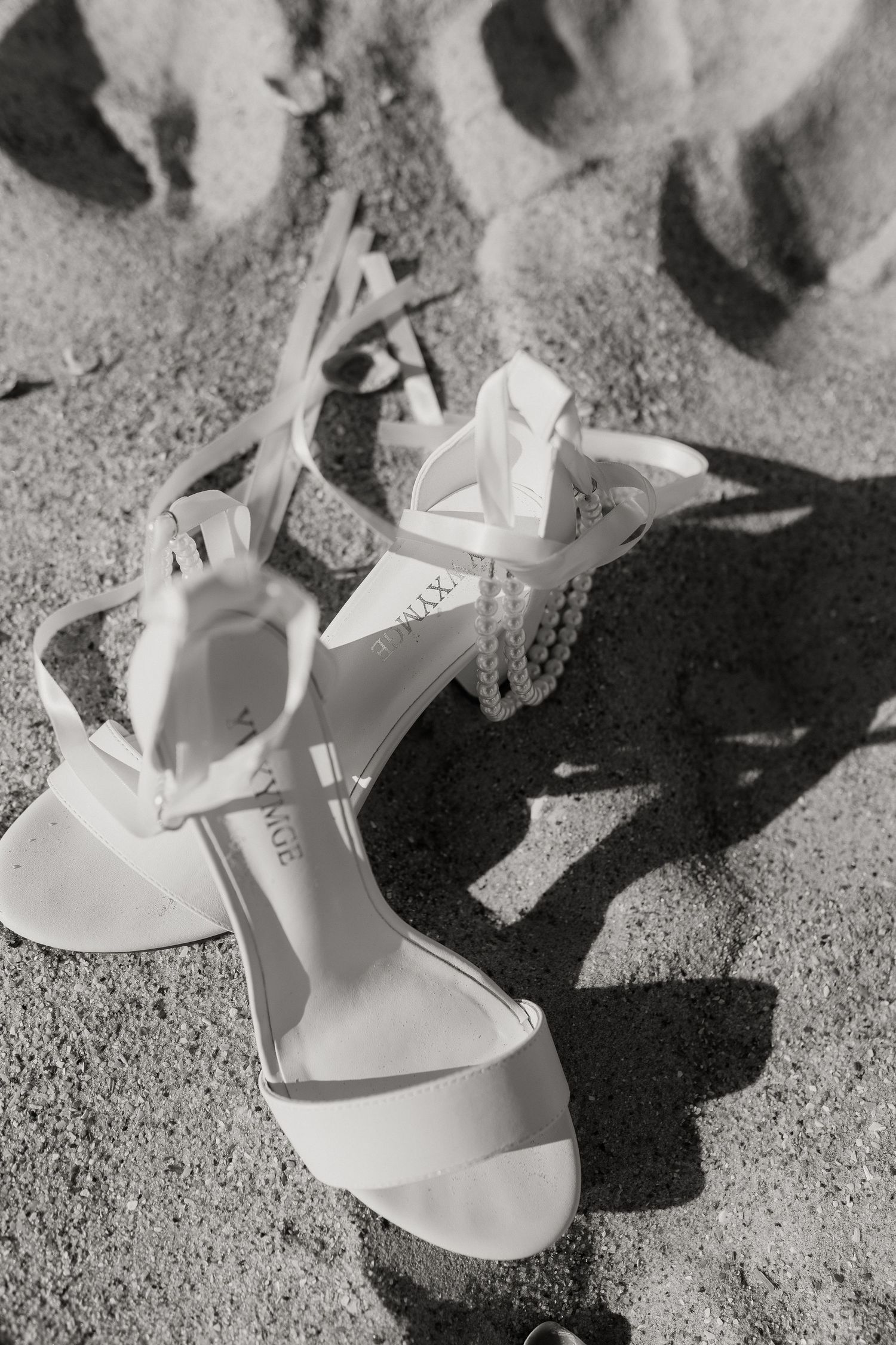 White wedding sandals with ribbon straps laying on sandy beach in black and white.