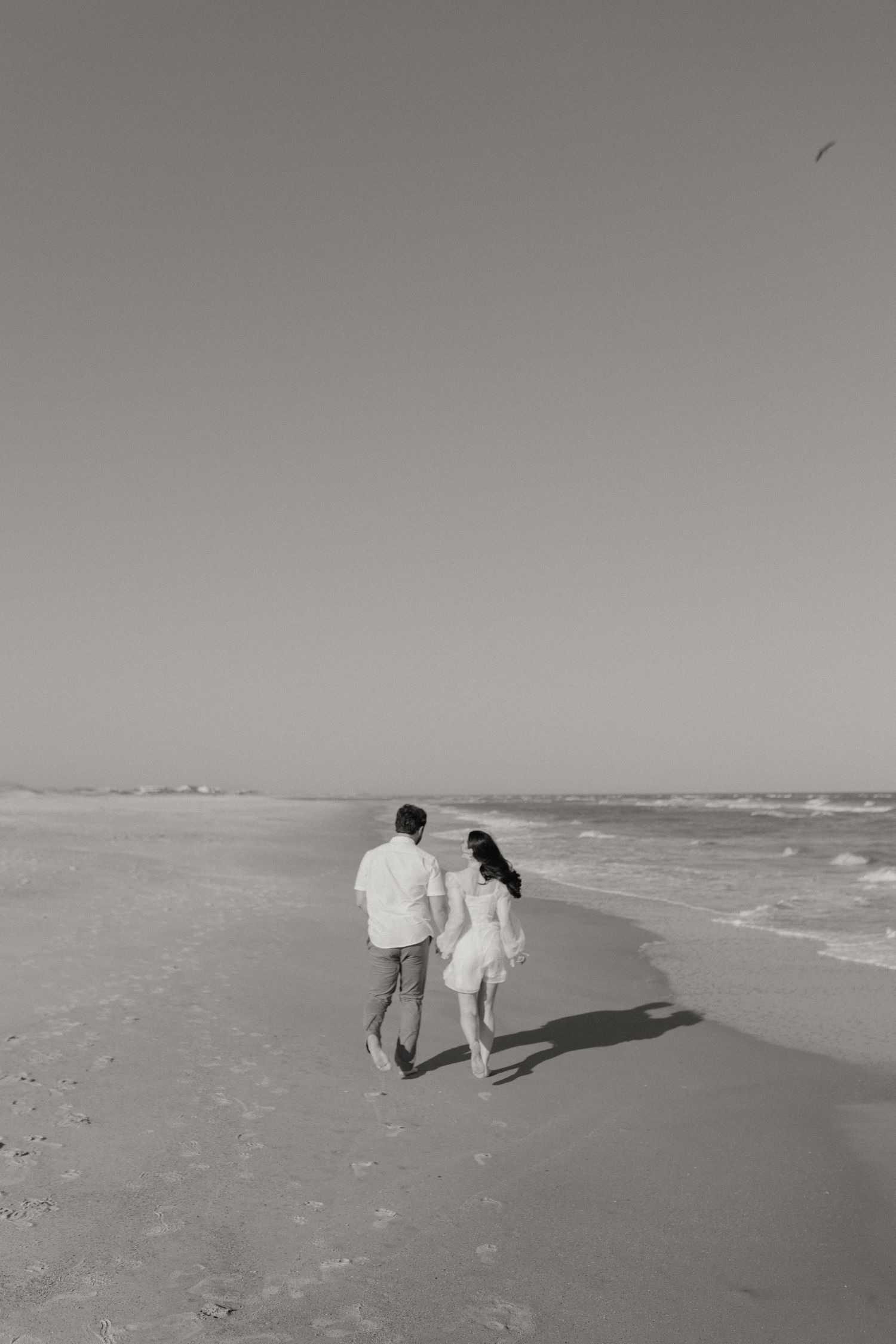 Couple walking hand in hand along a serene beach shoreline in black and white.