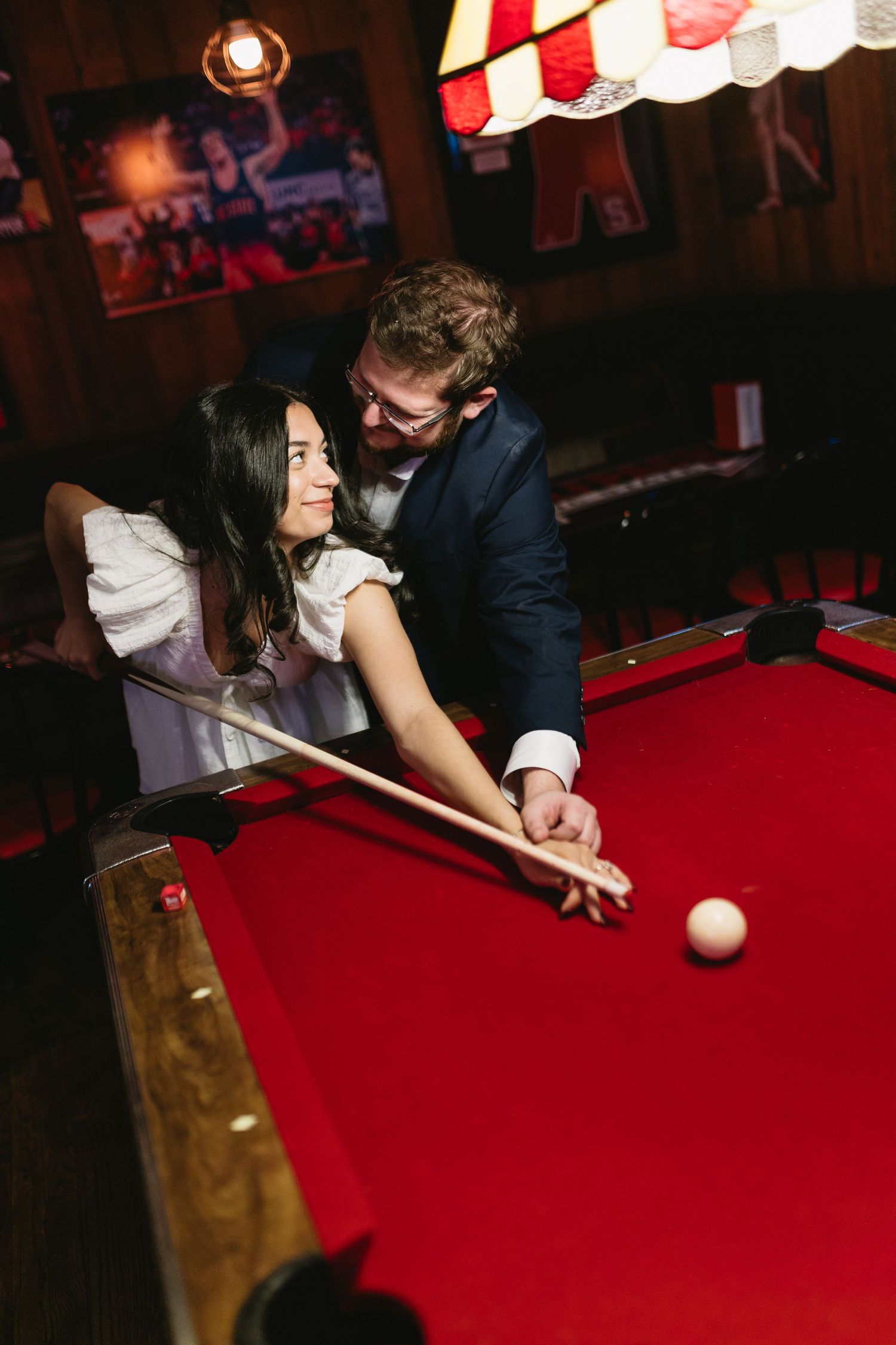 People playing pool on a red felt table in a bar setting with ambient lighting.
