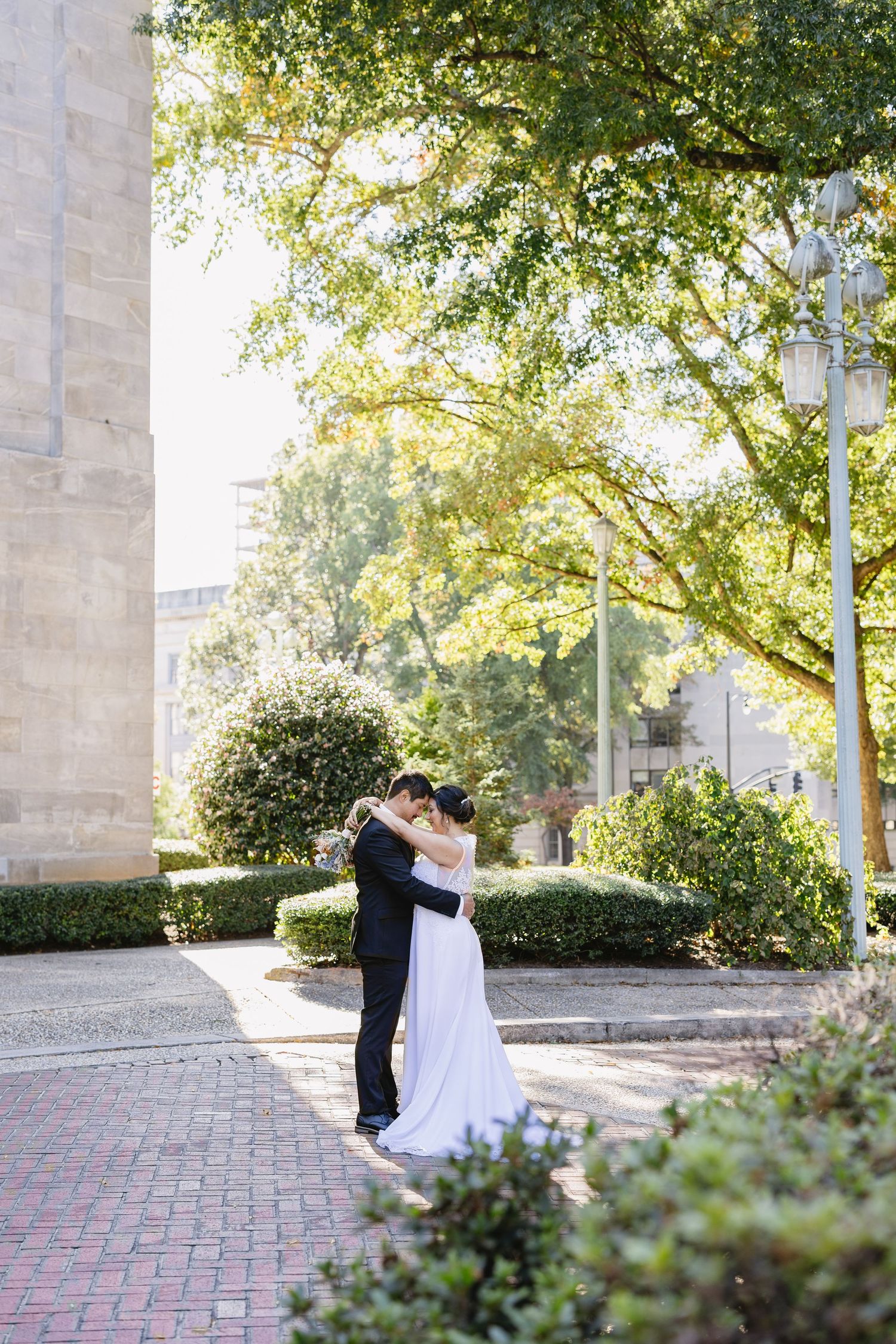 A couple dances in a sunlit courtyard surrounded by trees and brick pathways.