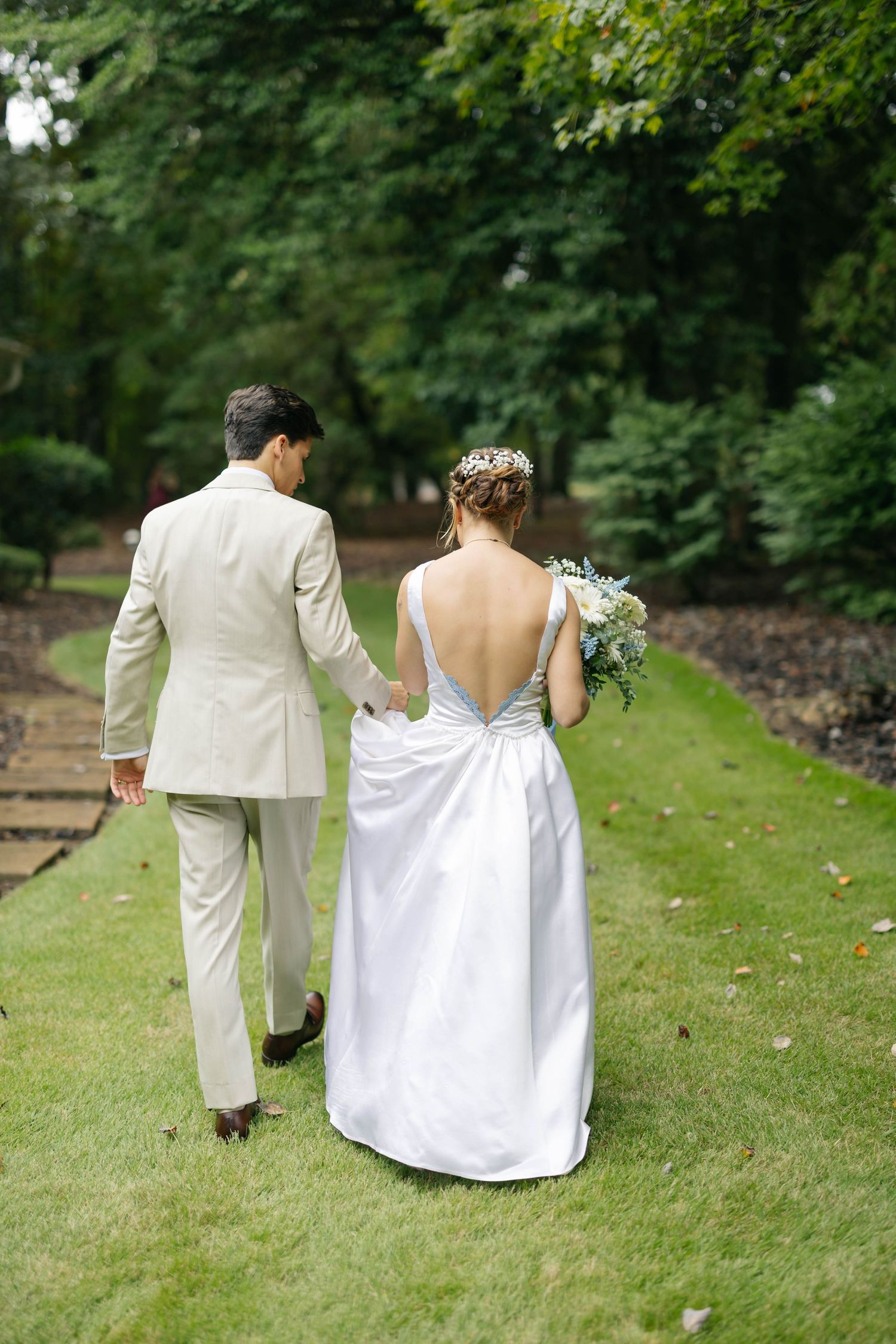 A couple walks together on a grassy path, the bride in white and groom in light-colored suit.