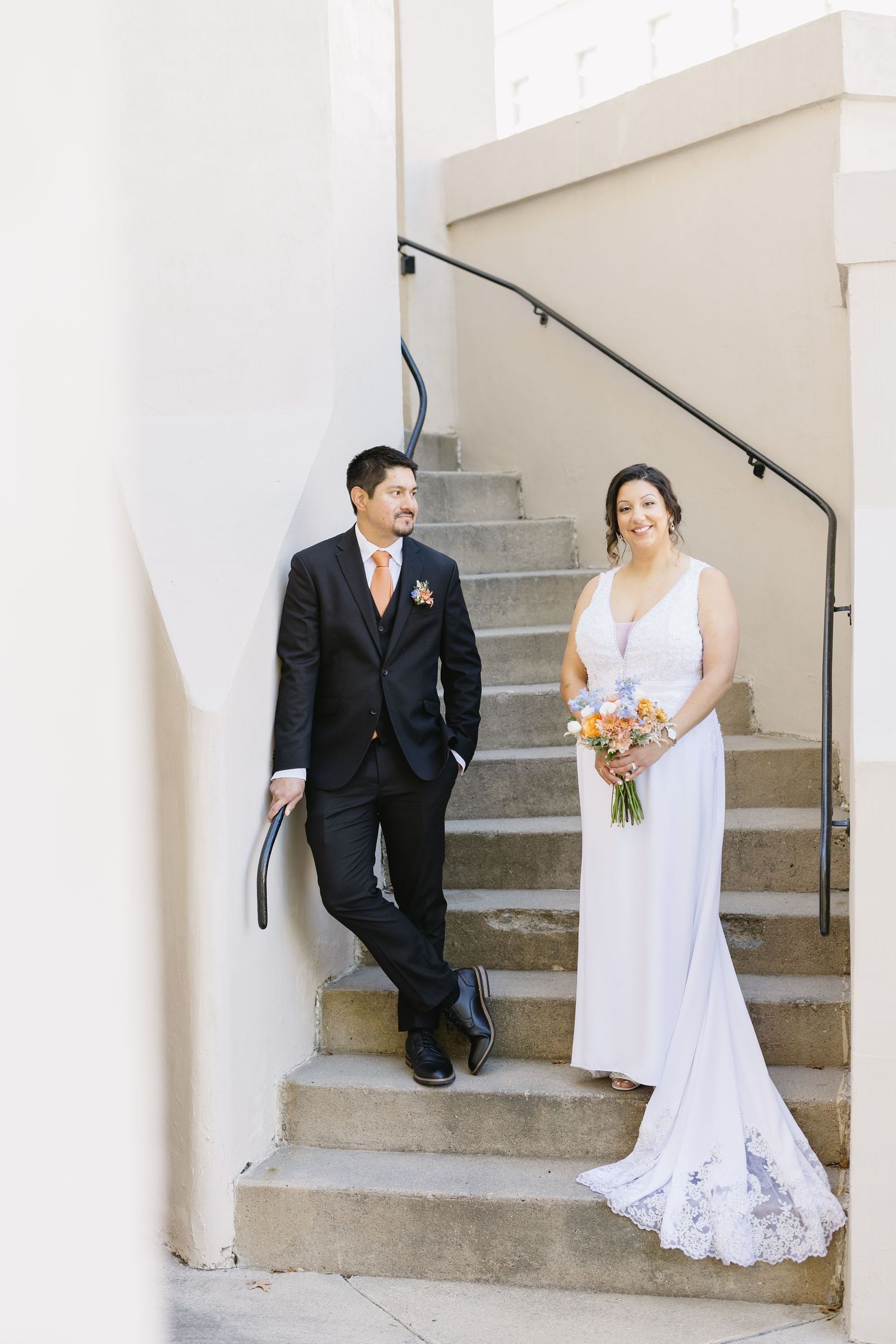 A couple shares a moment on concrete steps against a white wall with black handrails.
