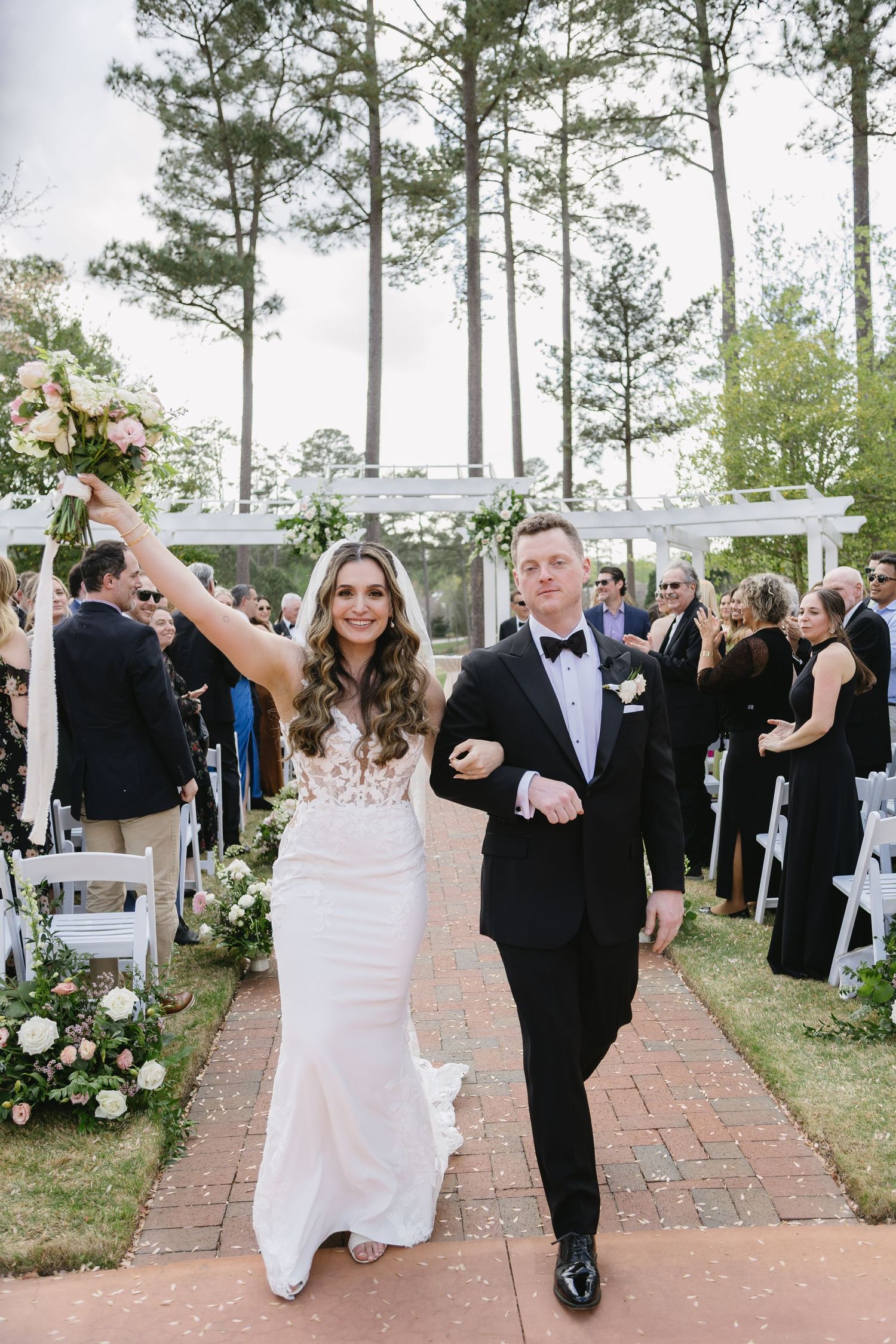 Joyful wedding ceremony procession on a brick path lined with tall pine trees.