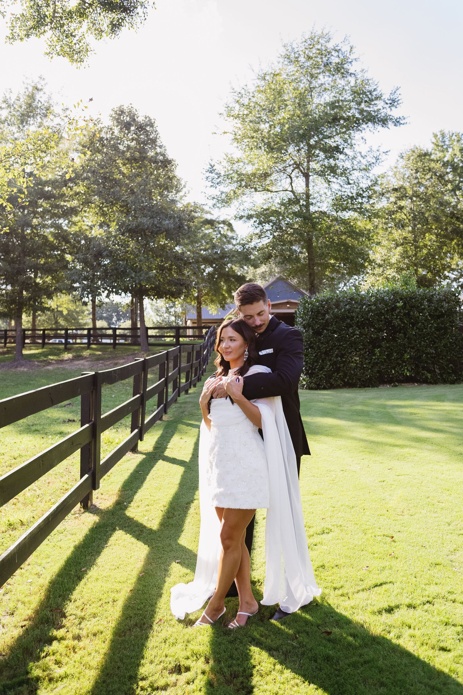A couple embraces by a wooden fence on a sunny lawn with long shadows.