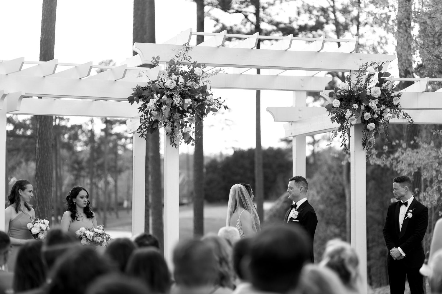 A wedding ceremony under a white pergola with floral arrangements.