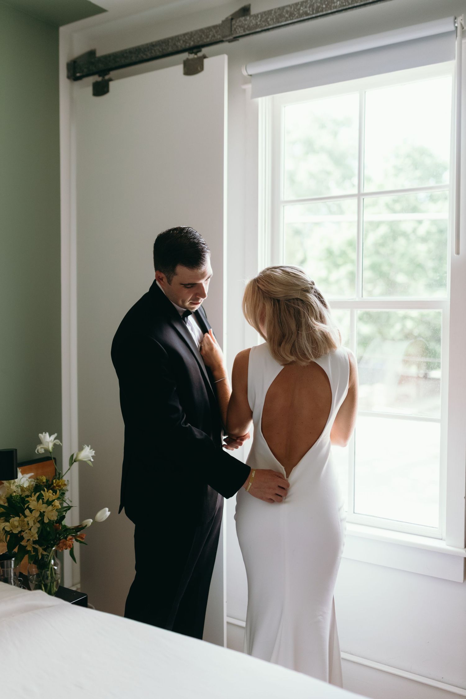 Bride and groom sharing intimate moment by window with floral arrangements.