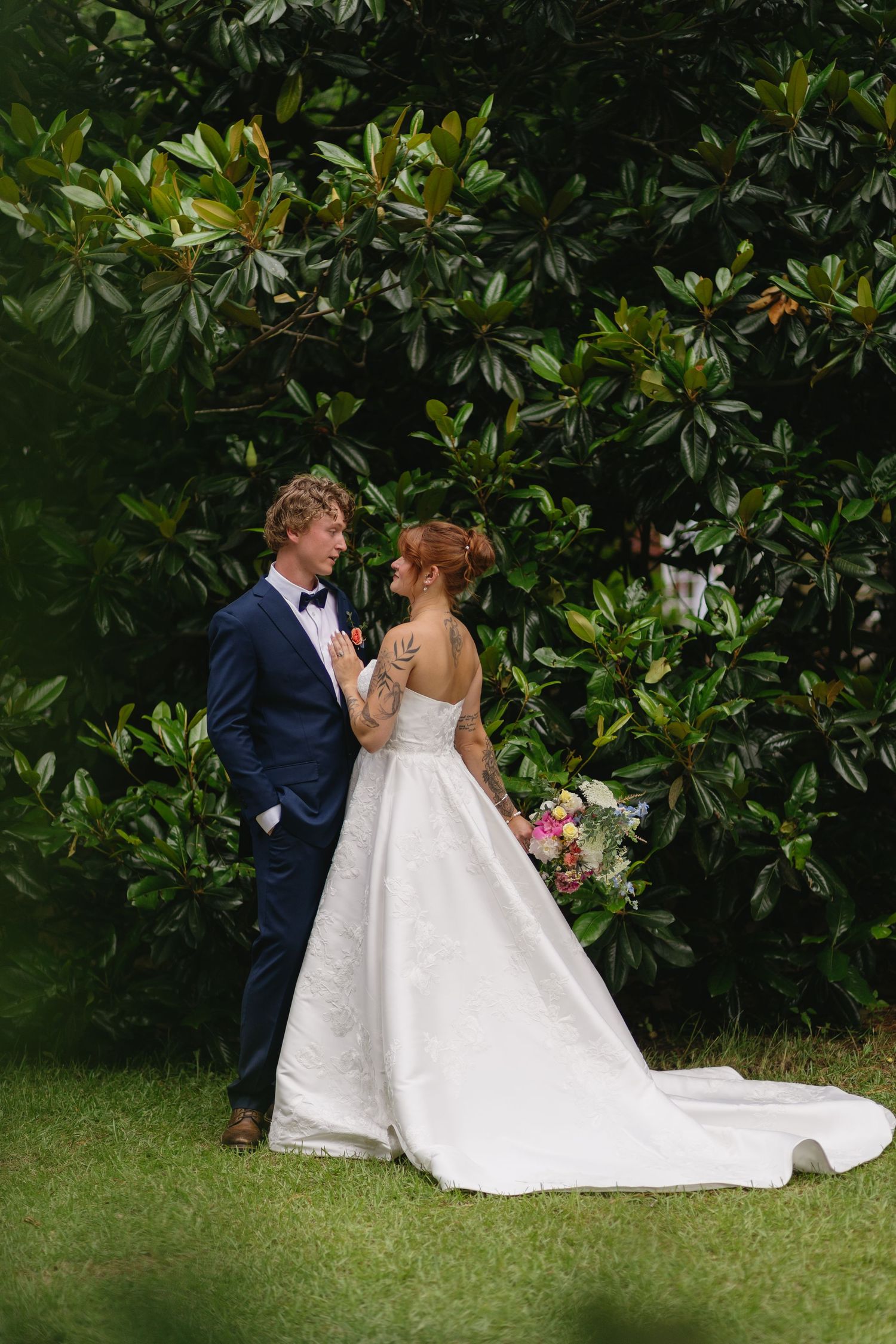 A couple poses against lush green foliage in formal wedding attire.