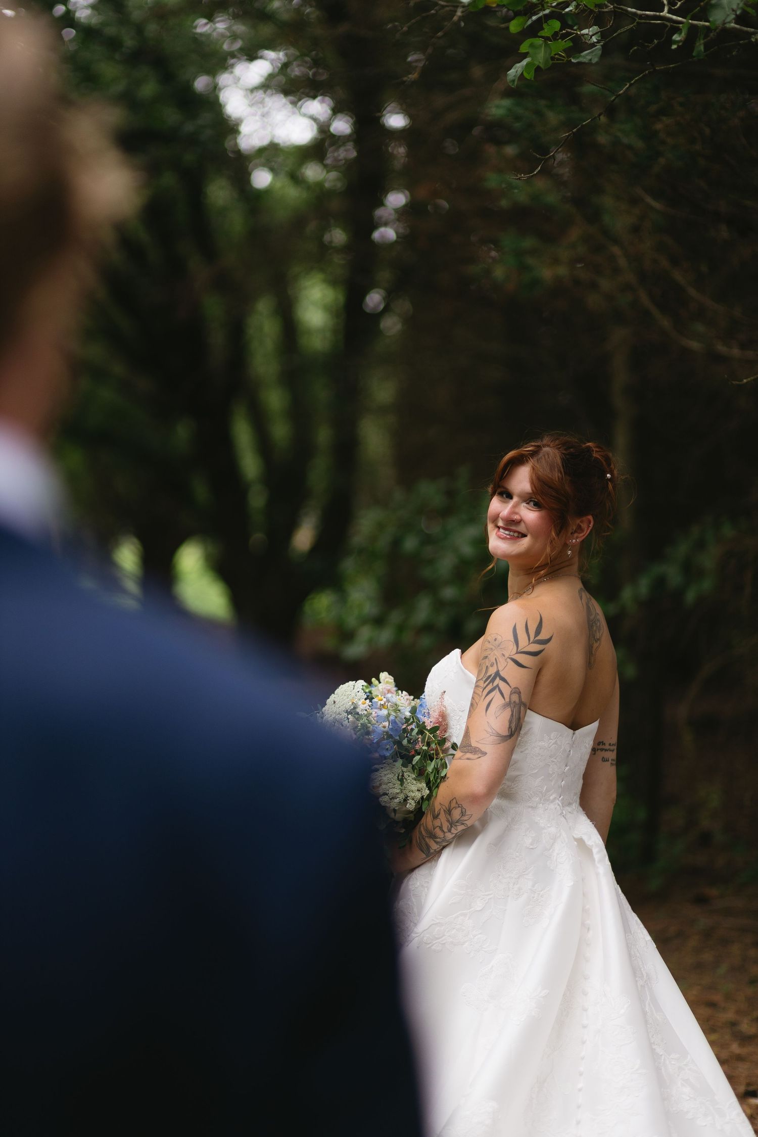 A bride in a white wedding dress smiles while holding a bouquet in a wooded outdoor setting.