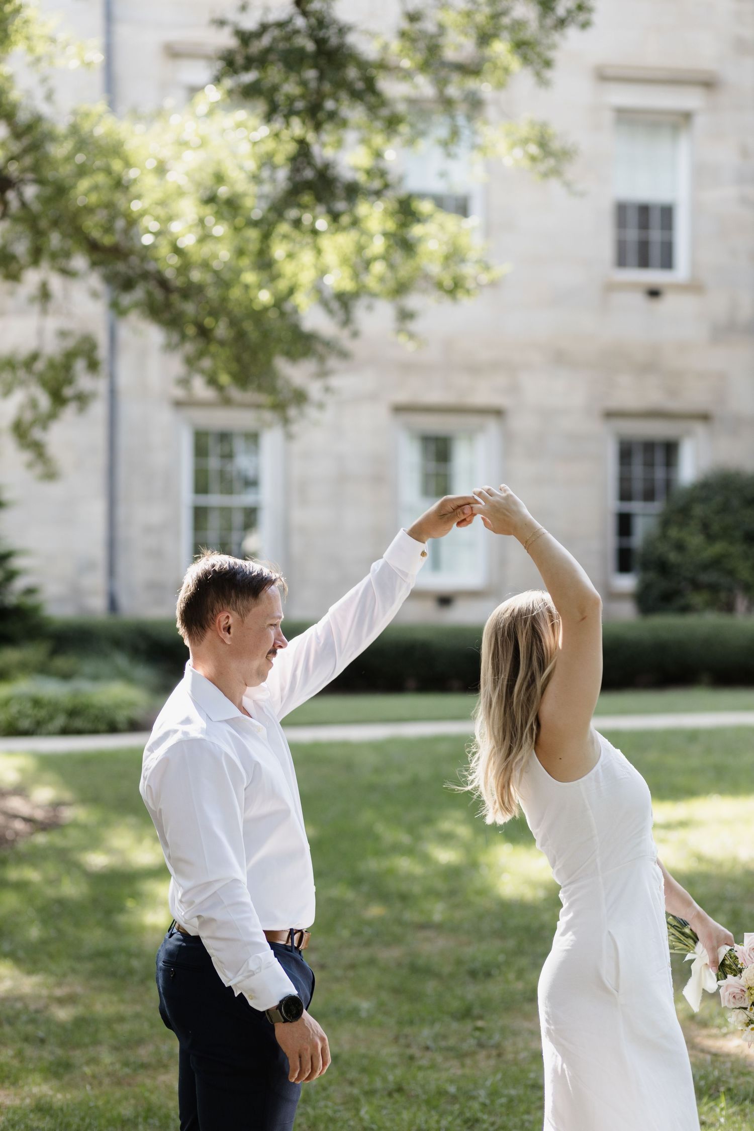 A couple dances in a garden setting with a historic building in the background.